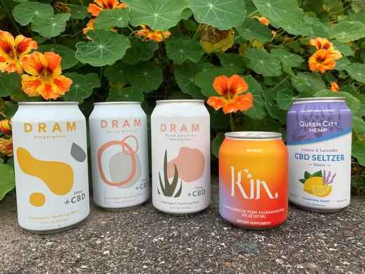 Cans of dram, kin, and queen city hemp beverages in a row inn front of flowers